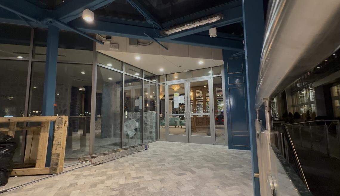 A high-angle, nighttime view of a restaurant entrance under construction. The scene features a modern glass-door entryway and large floor-to-ceiling windows framed in silver. Above the entrance is a dark blue structural overhang with recessed lighting and a space heater. In the foreground to the left, a wooden pallet and a silver ladder sit on a grey herringbone-patterned tile floor. Through the glass doors, the interior shows warm lighting, a chandelier, and a teal sofa.